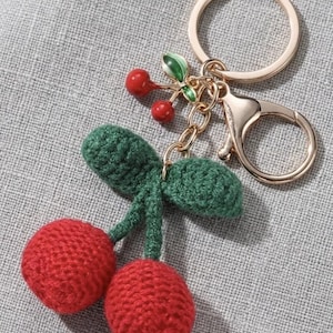 May include: A crochet cherry keyring in red and green, with a gold-coloured ring and clasp. The design includes two red cherries, green leaves, and a gold-coloured stem. The background is a grey fabric.