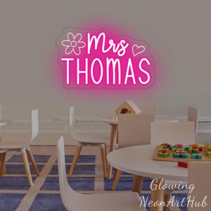 May include: A pink neon sign mounted on a wall displaying the text Mrs THOMAS in a cursive and sans-serif font. The sign features a line-art flower icon to the left of the text and a simple heart outline to the right, emitting a bright pink glow.