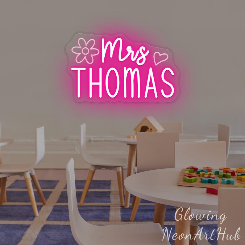May include: A pink neon sign mounted on a wall displaying the text Mrs THOMAS in a cursive and sans-serif font. The sign features a line-art flower icon to the left of the text and a simple heart outline to the right, emitting a bright pink glow.
