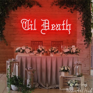 May include: A red neon sign saying "Til Death" is mounted on a wooden wall. The sign is above a table decorated with white floral arrangements and glassware. The table is draped with a light grey fabric. Decorative lanterns and greenery are placed around the table.