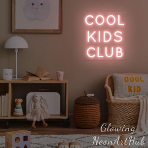 May include: A pink neon sign displaying "COOL KIDS CLUB" hangs in a child's room. The room features a wooden shelf, a lamp, and a woven chair. A cushion with the words "COOL KID" is on the chair, creating a playful atmosphere.