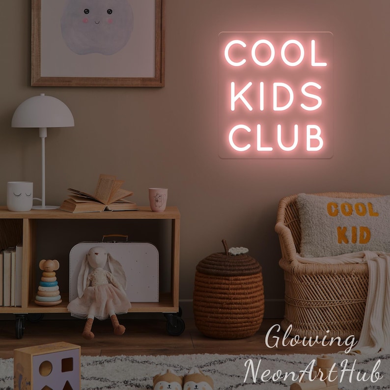May include: A pink neon sign displaying "COOL KIDS CLUB" hangs in a child's room. The room features a wooden shelf, a lamp, and a woven chair. A cushion with the words "COOL KID" is on the chair, creating a playful atmosphere.