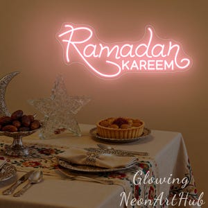May include: A pink neon sign that reads "Ramadan Kareem" is mounted on a wall. A table is set with a patterned tablecloth, silverware, and a tart. Decorative items include a crescent moon, a star, and a bowl of dates.