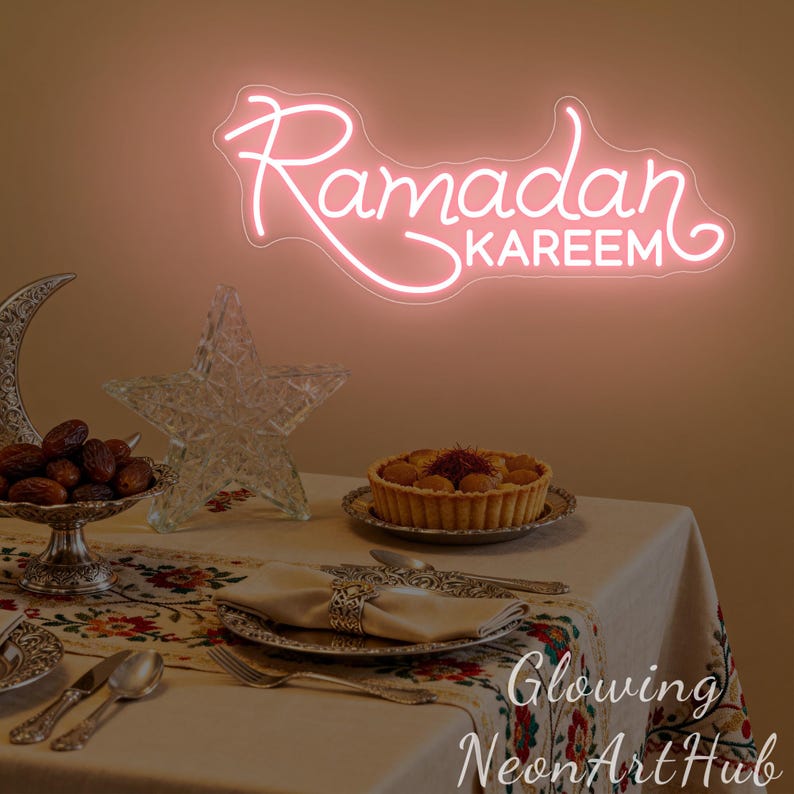 May include: A pink neon sign that reads "Ramadan Kareem" is mounted on a wall. A table is set with a patterned tablecloth, silverware, and a tart. Decorative items include a crescent moon, a star, and a bowl of dates.