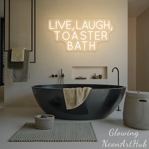 May include: A wall-mounted neon sign emitting a warm white glow, featuring the text LIVE, LAUGH, TOASTER BATH in a clean, sans-serif font. The sign is positioned above a large, matte black oval bathtub in a neutral-toned bathroom setting.
