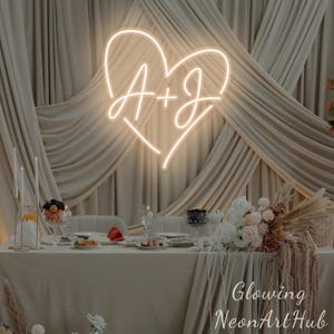 May include: A glowing neon sign in the shape of a heart with the letters "A + J" inside, hanging above a decorated table. The table is set for a special occasion, with flowers, candles, and food. The text "Glowing NeonArtHub" is visible.