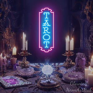 May include: A vertical neon sign with a pink border and the word TAROT in bright blue letters. The sign features a decorative scalloped edge and is displayed in a dark room with lit candles, a crystal ball, and various ornate objects.
