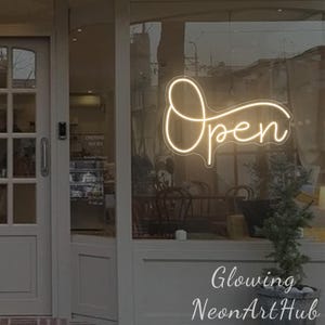May include: A glowing neon sign in a shop window displays the word "Open" in a cursive font. The sign is illuminated with a warm, inviting light. The shop's interior is visible through the glass, with tables and chairs. The text "Glowing NeonArtHub" is at the bottom.