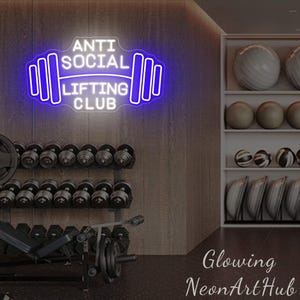 Pu&ograve; includere: Un'insegna al neon con la scritta "ANTI SOCIAL LIFTING CLUB" in bianco e blu, a forma di bilanciere. Lo sfondo mostra una palestra con manubri, dischi e palle da ginnastica.