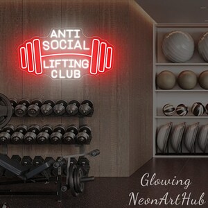 Pu&ograve; includere: Un'insegna al neon rossa e bianca con la scritta "ANTI SOCIAL LIFTING CLUB" in una palestra. L'insegna ha la forma di un bilanciere. Manubri e palle da ginnastica sono visibili sullo sfondo.