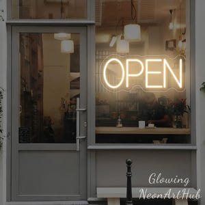 Pode incluir: Uma vitrine com uma placa de néon brilhante que diz "OPEN" em uma fonte cursiva. A placa é iluminada com uma luz quente e convidativa. O interior da loja é visível através da janela, com pessoas dentro. O nome da loja é "Glowing NeonArtHub".