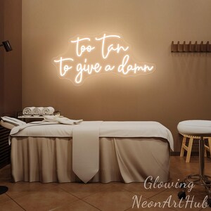 May include: A wall-mounted neon sign featuring the glowing white cursive text "too tan to give a damn" mounted on a brown wall above a spa treatment table covered with white linens and a white stool positioned to the right of the table.