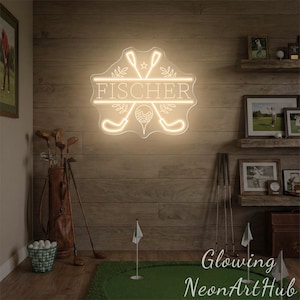 Puede incluir: Un letrero de ne&oacute;n con el nombre "FISCHER" en un marco decorativo, con palos de golf y una pelota de golf. El letrero se ilumina en un color blanco c&aacute;lido y est&aacute; montado en una pared de madera. Tambi&eacute;n se ven palos de golf y una bolsa de golf.