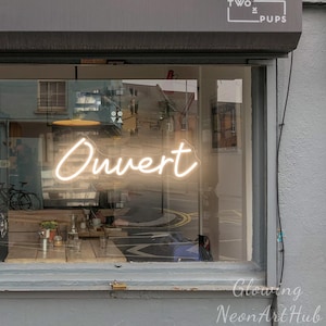 May include: A bright white neon sign displaying the word Ouvert in a cursive script font, mounted inside a storefront window. The sign is positioned above a wooden table and chairs, with the text Two Pups visible on the exterior awning above.