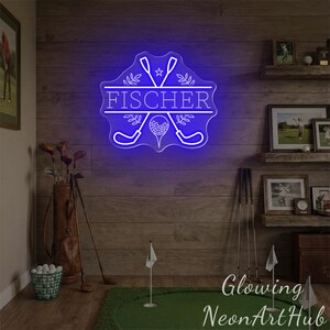 Puede incluir: Un letrero de ne&oacute;n azul brillante con el nombre "FISCHER" en blanco, con palos de golf, una pelota de golf y elementos decorativos. El letrero est&aacute; montado en una pared de madera, con decoraci&oacute;n tem&aacute;tica de golf.