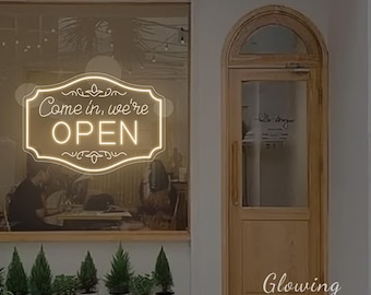 May include: A neon sign in a window displays the words "Come in, we're OPEN" in a decorative frame. The sign is illuminated with a warm, inviting glow. The shop's wooden door is to the right, and small potted plants line the window's base.