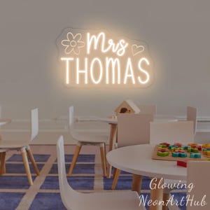 May include: A wall-mounted neon sign featuring the text Mrs THOMAS in a glowing warm white light. The design includes a simple outline of a flower to the left of the word Mrs and a small heart outline to the right, all set on a clear acrylic backing.