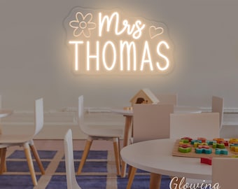 Puede incluir: Un letrero de neón montado en la pared con el texto Mrs THOMAS en una luz blanca cálida brillante. El diseño incluye el contorno de una flor a la izquierda de la palabra Mrs y un pequeño corazón a la derecha, sobre un soporte de acrílico.
