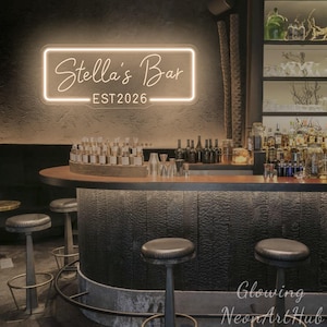 Può includere: Un'insegna al neon rettangolare montata su una parete scura e strutturata sopra un bancone da bar. L'insegna emana una luce calda e mostra la scritta Stella's Bar in corsivo, con il testo EST 2026 in un carattere sans-serif sottostante.
