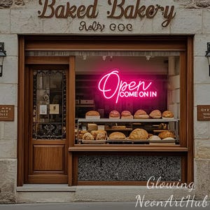 Puede incluir: Fachada de una panadería con puerta y escaparate de madera. Un letrero de neón en la ventana dice "Open COME ON IN" en rosa brillante. Varios panes y pasteles son visibles dentro de la vitrina. El nombre de la panadería, "Baked Bakery", se muestra encima de la entrada.