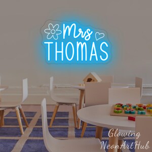May include: A bright blue neon sign mounted on a wall featuring the text Mrs THOMAS in a cursive and sans-serif font, accompanied by a line-art flower icon on the left and a small heart icon on the right, emitting a soft glow against the wall.