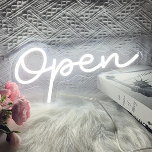 May include: A white neon sign in cursive script that reads "Open". The sign is illuminated and sits on a fluffy white surface. A book with the Statue of Liberty on the cover is visible in the background, along with pink flowers.