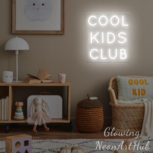 May include: A white neon sign displaying "COOL KIDS CLUB" on a beige wall. The room is furnished with children's items, including a lamp, a woven chair, and a pillow with "COOL KID" written on it. The overall theme is a children's room.