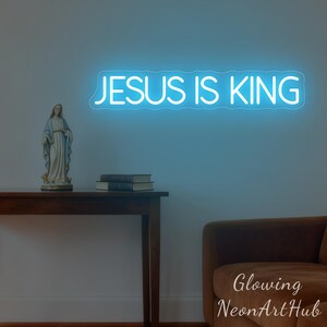 May include: A blue neon sign that reads "JESUS IS KING" is mounted on a wall. A statue of a religious figure and two books sit on a wooden table. A brown sofa is in the lower right corner.