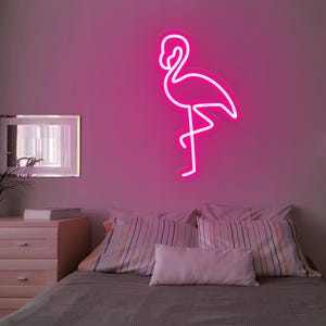 Puede incluir: Un letrero de neón rosa brillante con forma de flamenco sobre una pared rosa. El flamenco tiene un contorno blanco. El letrero está encima de una cama con almohadas a rayas y una colcha gris. Un espejo y una cómoda son visibles.