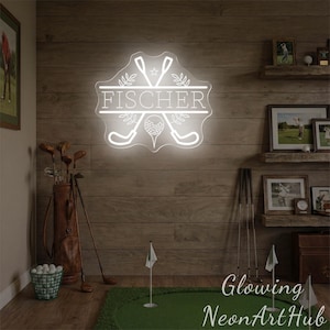 Puede incluir: Un letrero de ne&oacute;n LED blanco montado en una pared con paneles de madera, que presenta el nombre FISCHER en letras may&uacute;sculas, flanqueado por palos de golf cruzados, una pelota de golf y motivos de hojas, dentro de un borde decorativo.