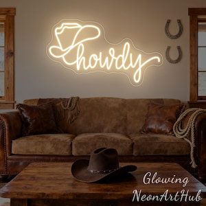 以下が含まれることがあります： カウボーイハットの形をしたネオンサインと、筆記体で「Howdy」という言葉。 サインは茶色の革張りのソファの上にある壁に取り付けられています。 壁には2つの蹄鉄があります。 茶色のカウボーイハットが木製のコーヒーテーブルの上にあります。