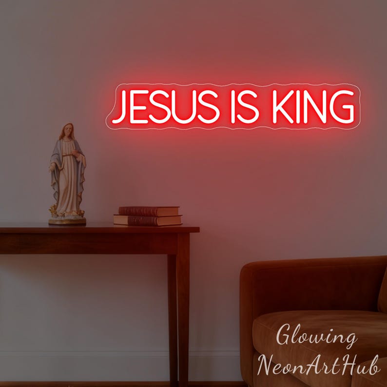 May include: A red neon sign displaying the text "JESUS IS KING" is mounted on a wall. A statue and books sit on a wooden table below the sign. A brown sofa is in the lower right corner. The sign is from Glowing NeonArtHub.
