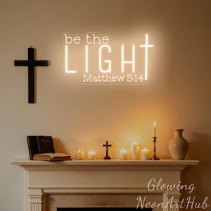 May include: A glowing neon sign on a wall displays the text be the LIGHT Matthew 5:14 with a cross integrated into the letter T. To the left hangs a black wooden cross. Below, a mantel holds books, a small cross, candles, and a vase.