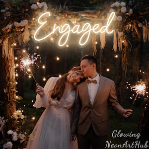 Puede incluir: Una escena romántica con una pareja sosteniendo bengalas bajo un letrero de neón que dice "Engaged". El escenario es al aire libre, decorado con luces de hadas y arreglos florales. El texto "Glowing NeonArtHub" es visible en la parte inferior de la imagen.