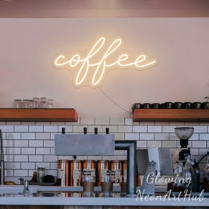Könnte beinhalten: Ein leuchtendes Neon-Schild mit dem Wort "coffee" in Schreibschrift an einer Wand. Das Schild ist warmweiß. Der Hintergrund zeigt eine Kaffeehaus-Theke mit Geräten und Regalen.