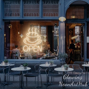 以下が含まれることがあります： カフェの店頭。窓にはネオンサインがあり、コーヒーカップのデザインと「OPEN」の文字が表示されています。屋外にはテーブルと椅子があり、カフェの営業を示唆しています。