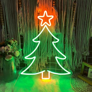 Könnte beinhalten: Ein Neon-Weihnachtsbaumlicht mit einem Stern oben. Der Baum ist in hellem Grün umrandet, mit einem orangefarbenen Stern und Sockel. Der Hintergrund zeigt einen Makramee-Wandbehang und eine Blumenvase.