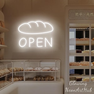 May include: A white neon sign in the shape of a loaf of bread with the word "OPEN" below it. The sign is mounted on a light-colored wall. A glass display case filled with baked goods is below the sign.