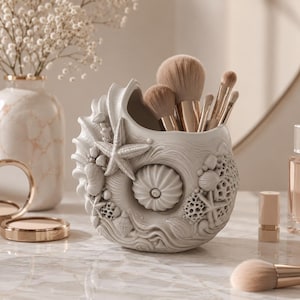 May include: A white seashell-shaped makeup brush holder with a starfish and other sea life details. The holder contains several makeup brushes with light brown bristles. The holder is on a marble surface.