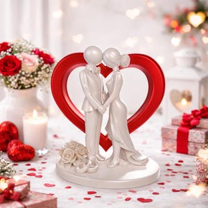 Può includere: Un cake topper bianco per matrimonio con una figura di sposi che si tengono per mano, ambientato su uno sfondo a forma di cuore rosso. Le figure sono vestite in abiti formali, con un bouquet di rose bianche alla base. La scena è decorata con coriandoli a forma di cuore rosso.