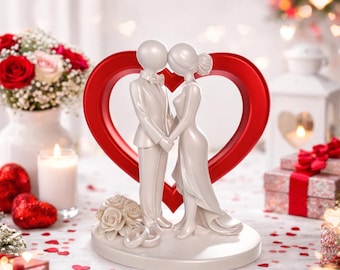 May include: A white wedding cake topper featuring a bride and groom figure holding hands, set against a red heart-shaped backdrop. The figures are dressed in formal attire, with a bouquet of white roses at the base. The scene is decorated with red heart confetti.