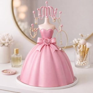 Può includere: Un organizer per gioielli rosa a forma di manichino. Il manichino ha un fiocco e un dettaglio a forma di rosa in vita e una parte superiore decorativa con ganci per appendere i gioielli. Diversi gioielli sono esposti sull'organizer.