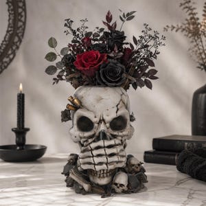 May include: A white skull-shaped vase with a cracked finish, adorned with faux bullet casings and small skulls at the base. The vase holds a bouquet of red and black roses and dark foliage, creating a gothic decorative piece.