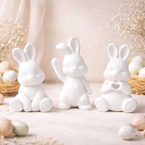 May include: Three white ceramic rabbit figurines in different poses. One sits with legs crossed, another waves, and the third forms a heart shape with its hands. Easter decorations.