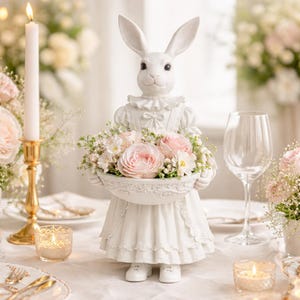 Puede incluir: Una figura de conejo de cerámica blanca, vestida con un vestido con volantes, sostiene una cesta de flores rosas y blancas. El conejo tiene orejas largas y está posado sobre una mesa decorada con velas y cristalería.