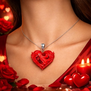 Puede incluir: Un collar de plata con un colgante de corazón rojo. El colgante de corazón tiene un diseño tallado y texturizado. El collar se usa sobre un fondo rojo, con velas y pétalos de rosa visibles.