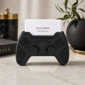 以下が含まれることがあります： ビデオゲームコントローラーの形をしたマットブラックの名刺ホルダー。ホルダーには方向パッド、2つのアナログスティック、4つのアクションボタンが付いています。中央には「Your Name Your Business」というテキストが書かれた白い名刺が差し込まれています。