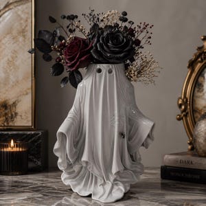 Peut inclure: Un vase blanc en céramique en forme de fantôme contenant des roses rouges et noires foncées et des fleurs séchées. Le vase a deux trous pour les yeux et un design drapé. Une araignée et une toile sont sur le côté. Le vase est sur une surface en marbre.