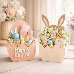 Puede incluir: Dos recipientes decorativos con forma de huevo con temática de Pascua. Uno es de color melocotón con el texto "Happy Easter", un conejito azul y huevos. El otro es de color crema con orejas de conejo, flores y un conejito dorado. Ambos contienen regalos.