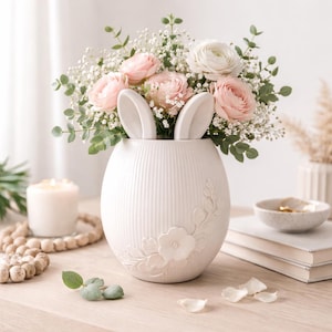 Può includere: Un vaso in ceramica bianca a forma di uovo, con orecchie da coniglio e dettagli floreali. Il vaso contiene un bouquet di rose rosa e bianche, velo da sposa e verde, un centrotavola a tema primaverile.